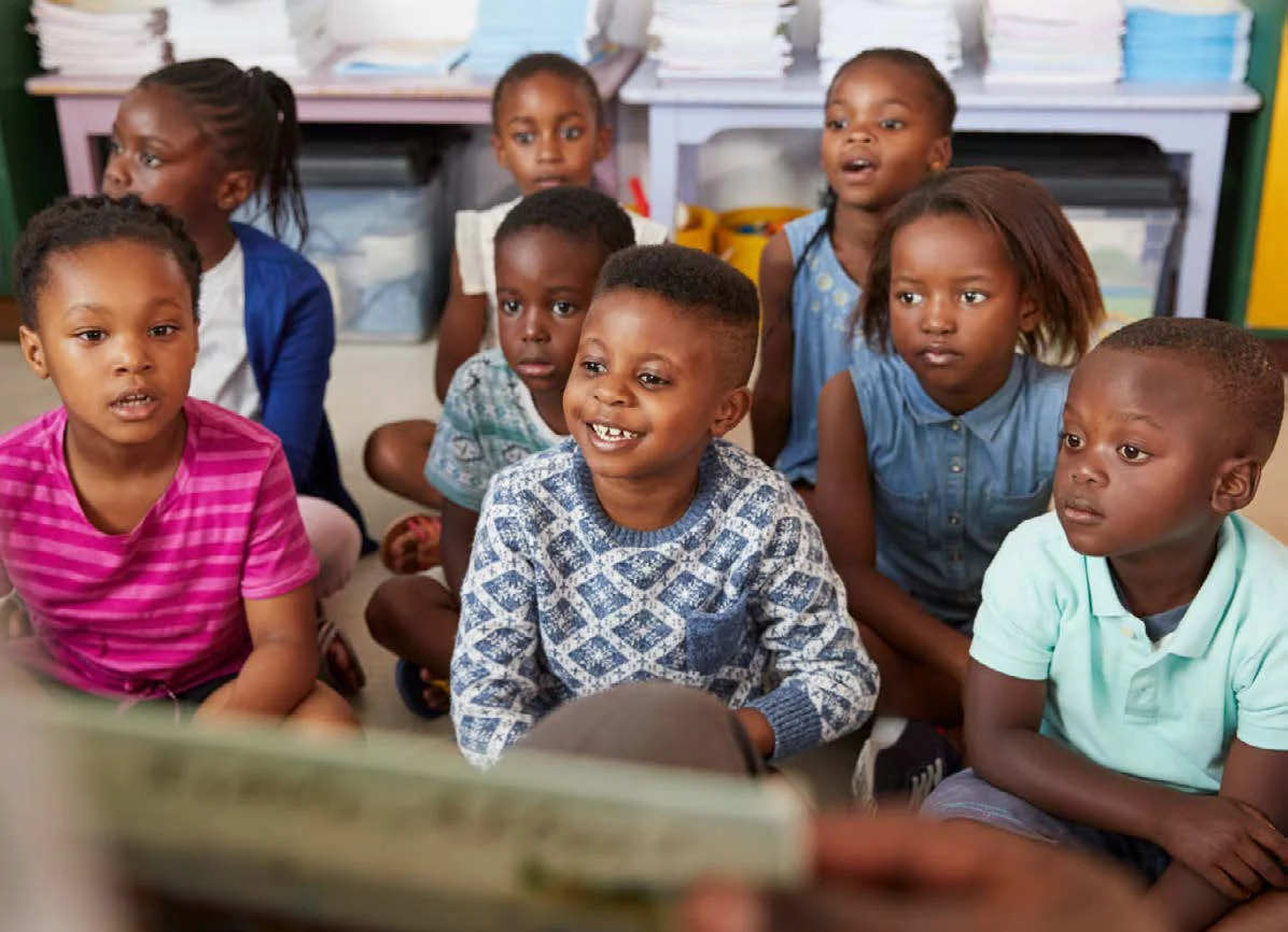 A group of young children sit together on a classroom carpet, looking up and smiling.