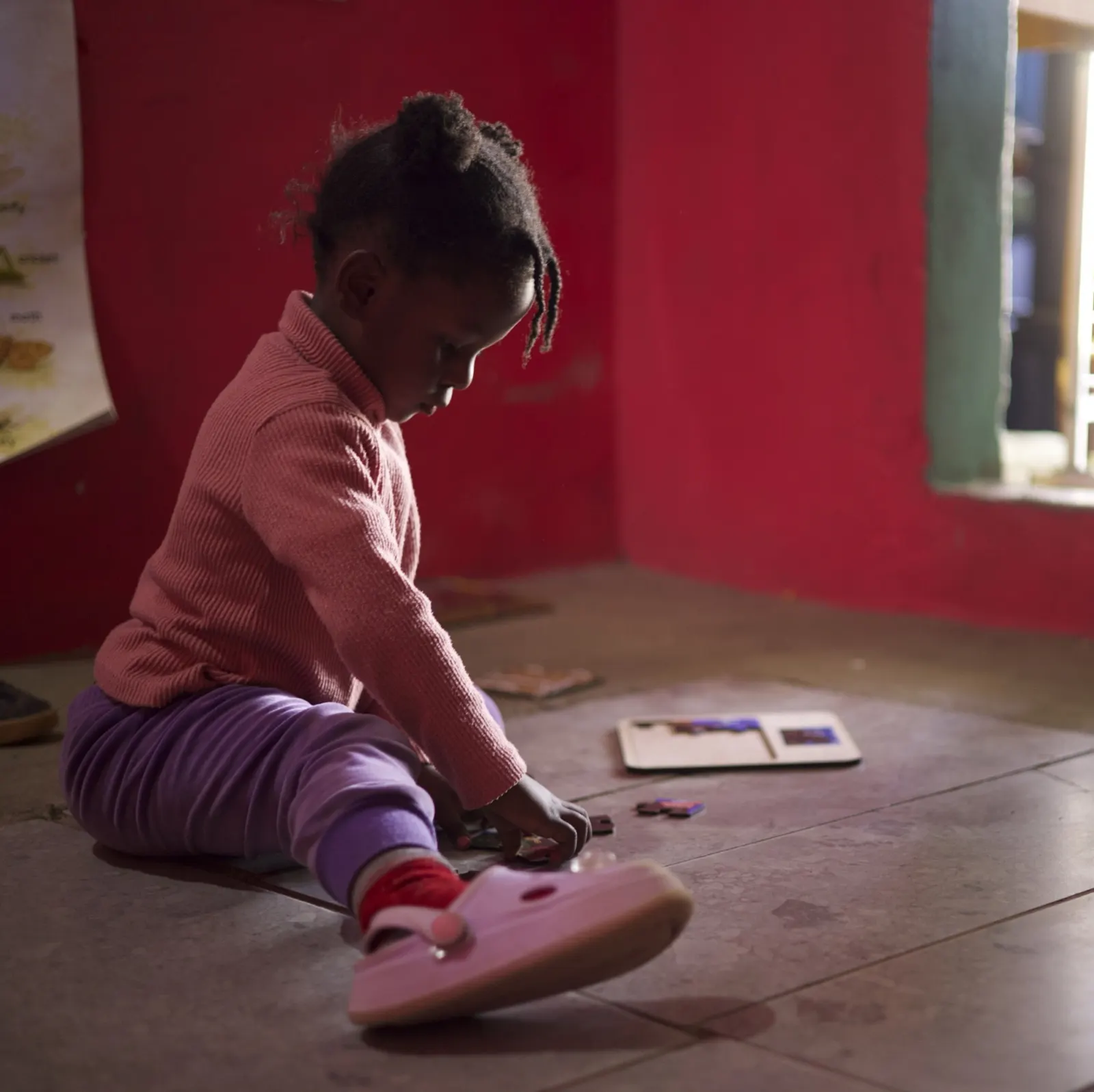 A young girl in a pink top plays on the floor of a dim classroom.