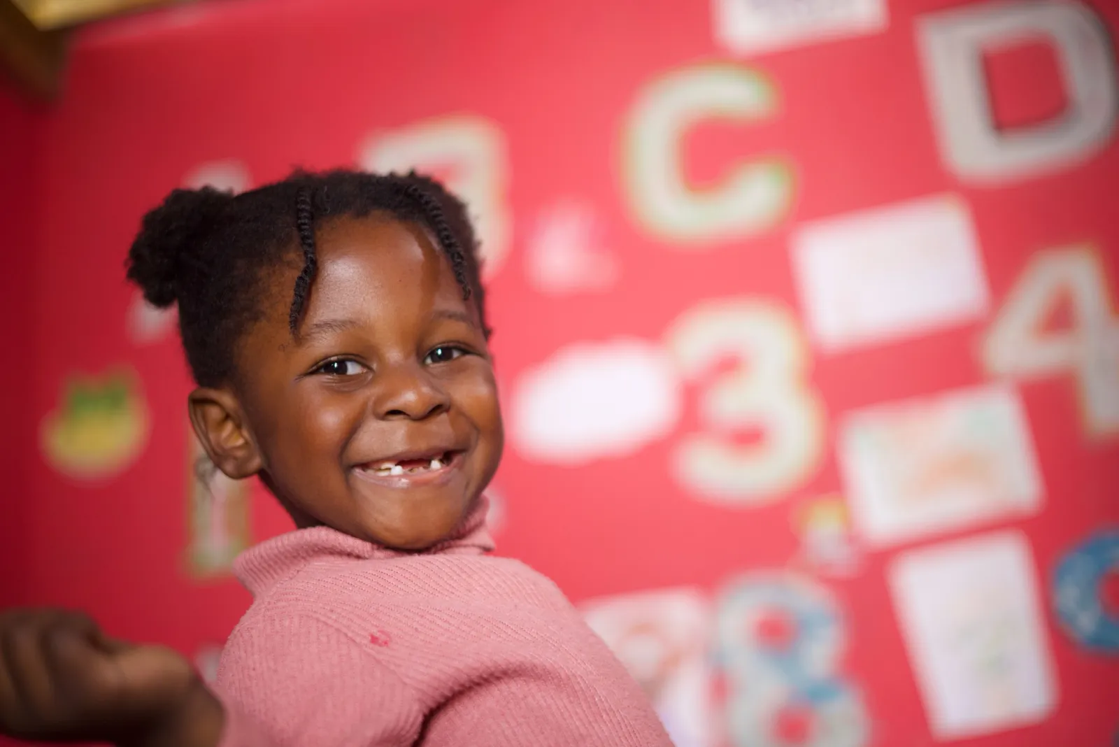 A young girl with braided hair, wearing a pink jersey, grins at the camera.