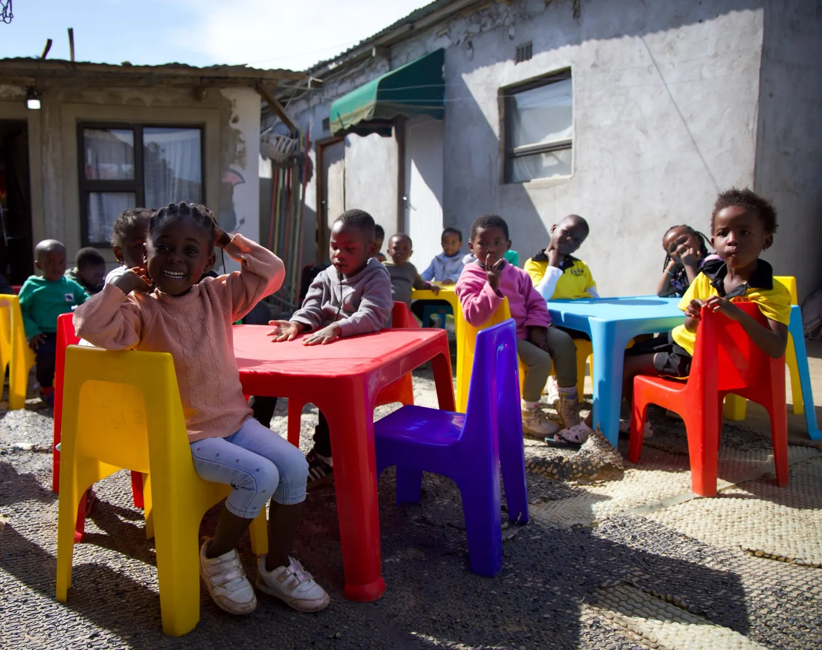 Children sit on colourful plastic chairs at red and blue tables in a yard.
