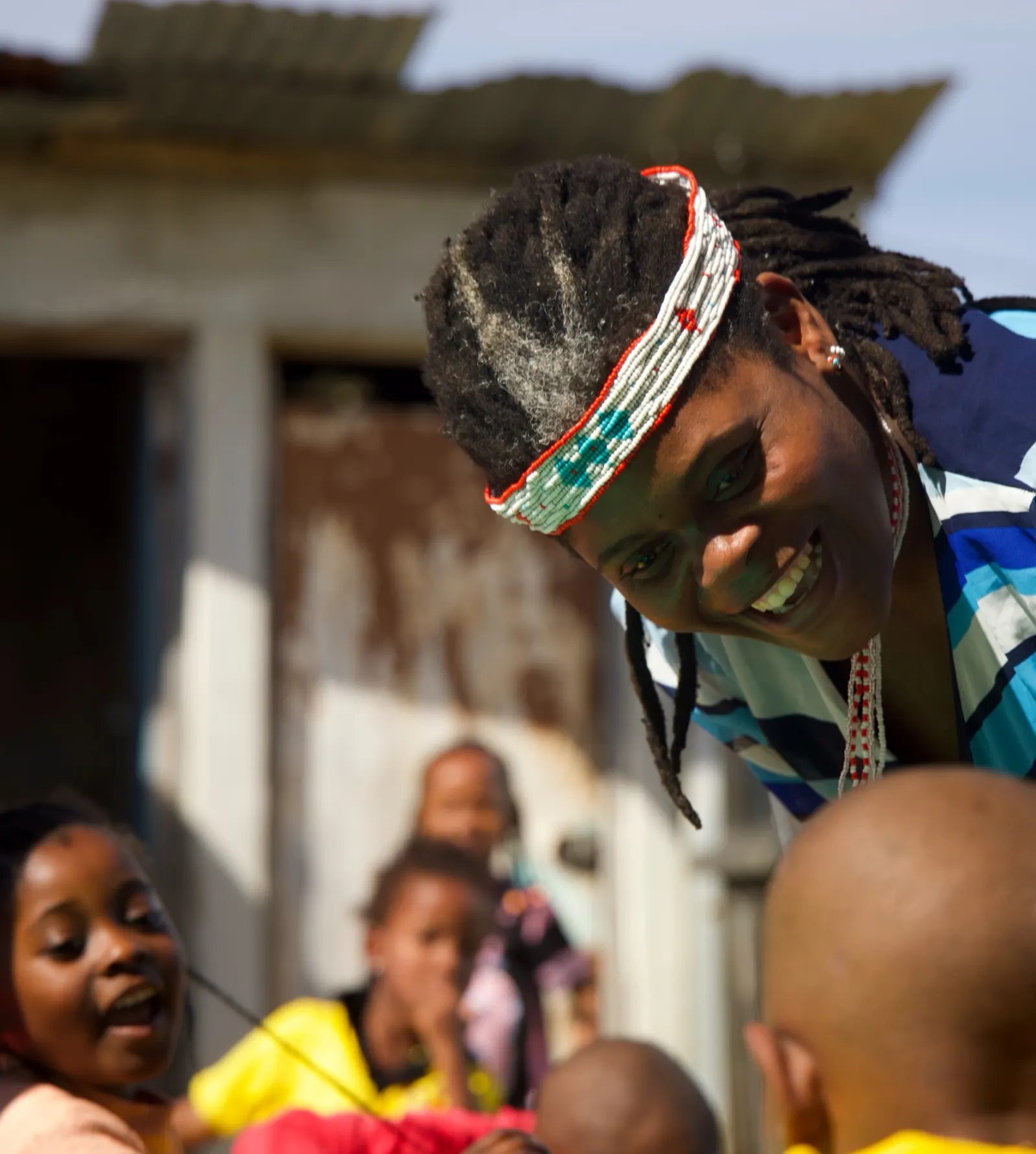 Andiswa Damane, wearing a beaded headband, leans down smiling towards a group of children.