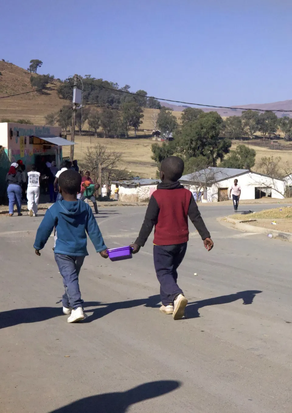 Two young children walking hand in hand down a road towards a creche, one carrying a lunch box.